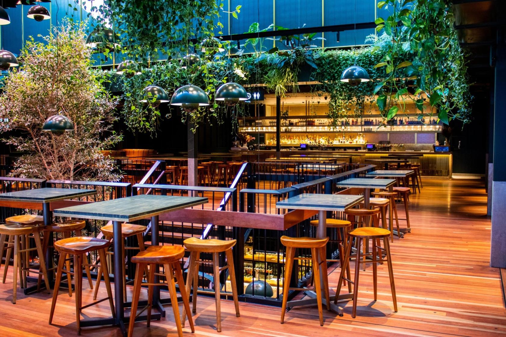 An event space in a bar with wooden tables, high stools, hanging plants, and a visible bar counter at The Sound Garden.