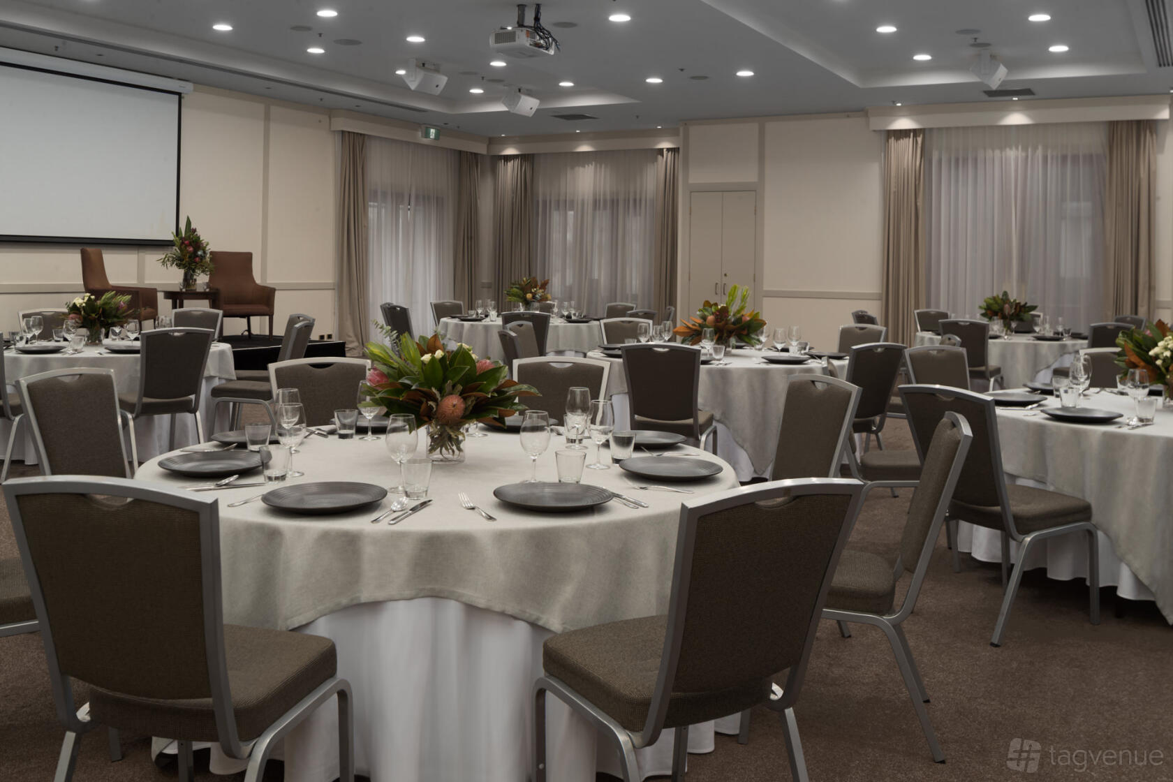 An event space with round tables set with neutral linens, floral centerpieces, and a stage at Yarra Valley Lodge.