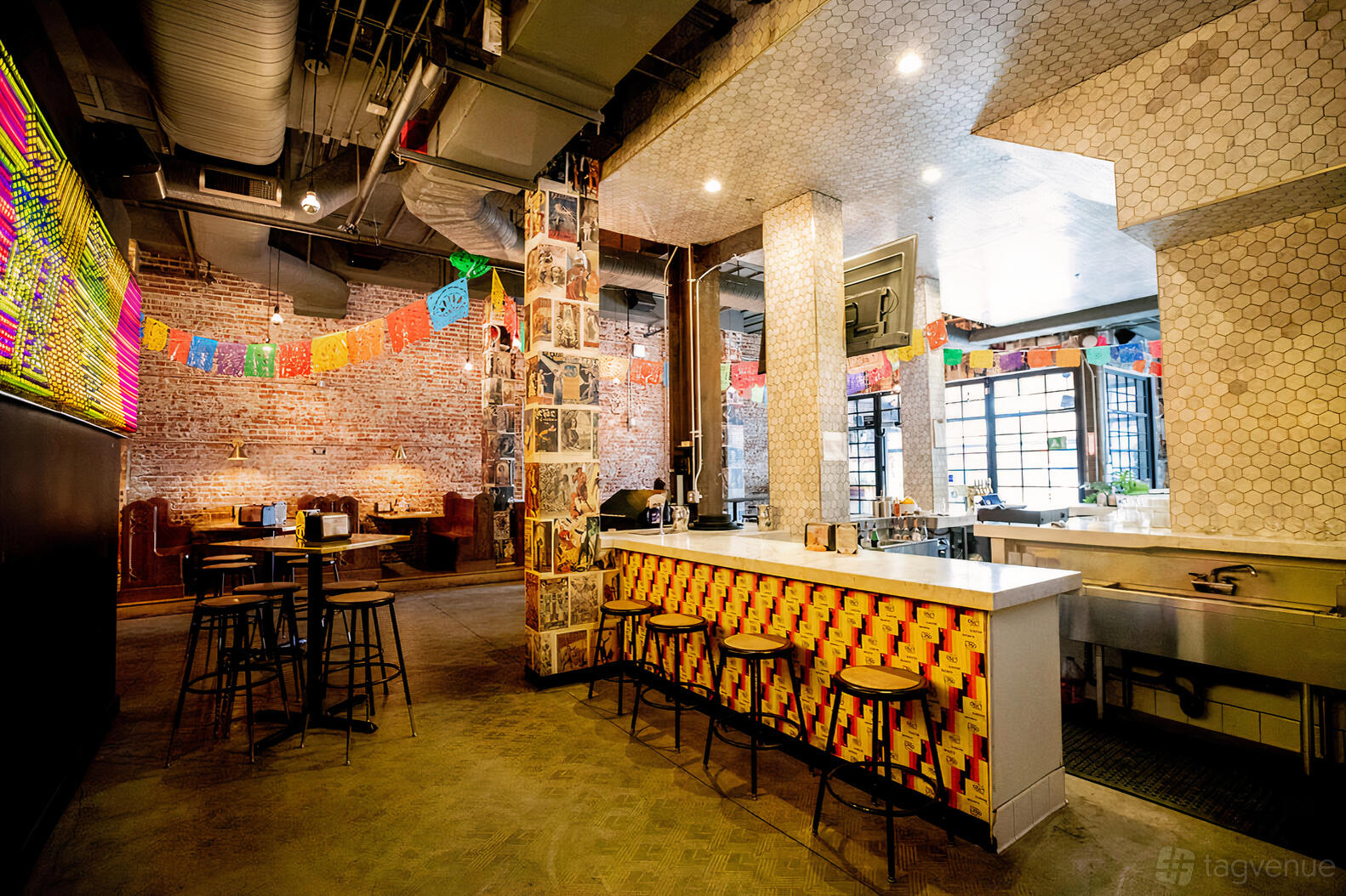A bar with exposed brick walls, colorful papel picado banners, and a counter with bar stools at Native Son LA.