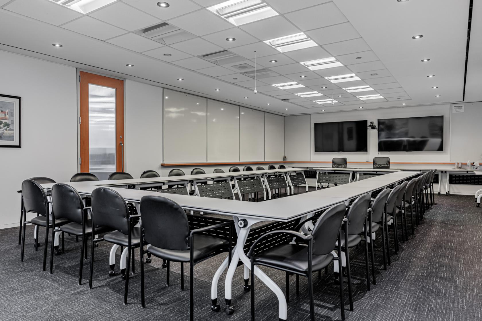 A meeting room with U-shaped tables, black chairs, dual wall-mounted screens, and ceiling lights at The Rostie Group.