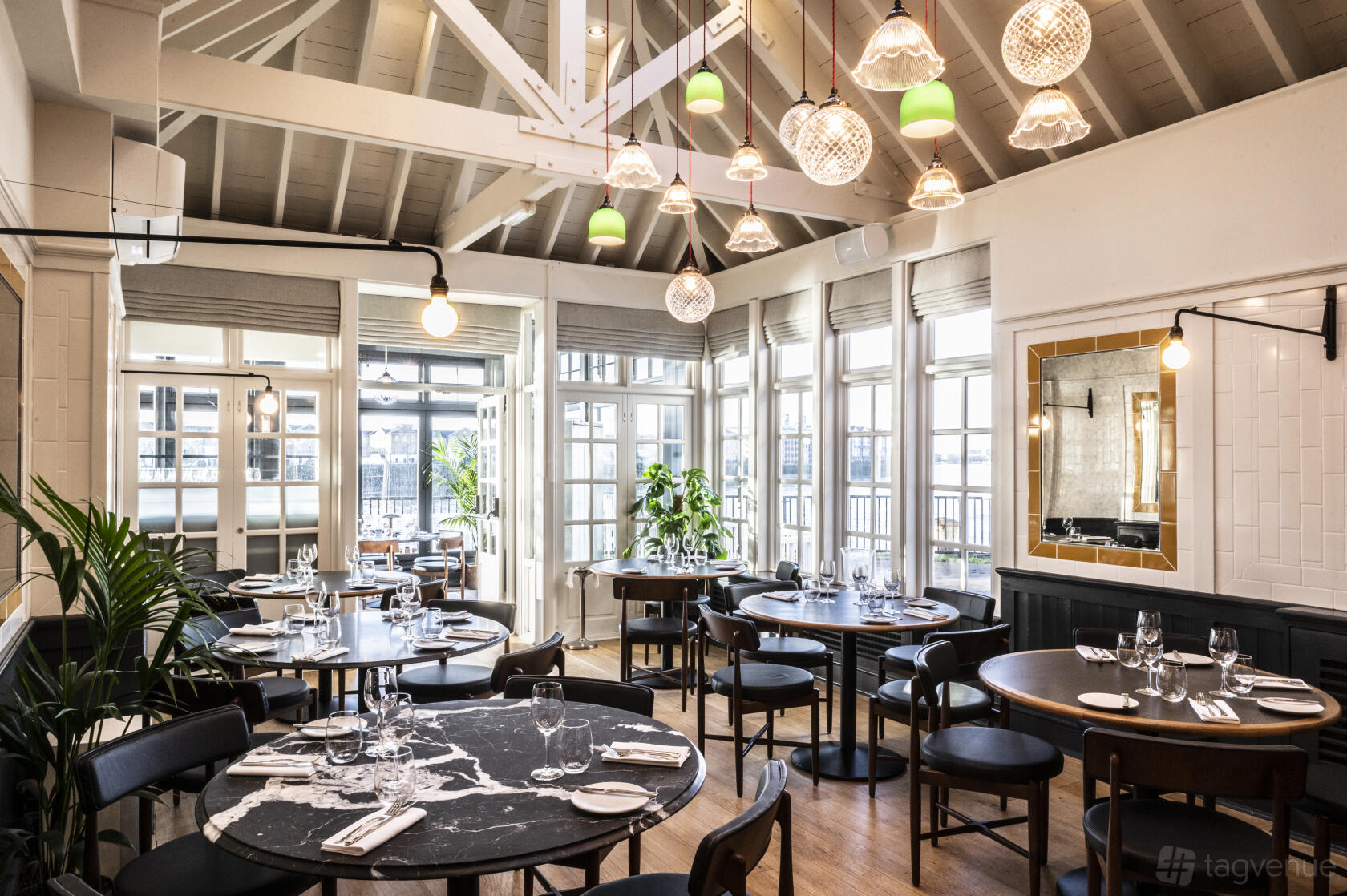 A dining room with round black tables, wooden flooring, large windows, and pendant lighting at Bread Street Kitchen, Limehouse.