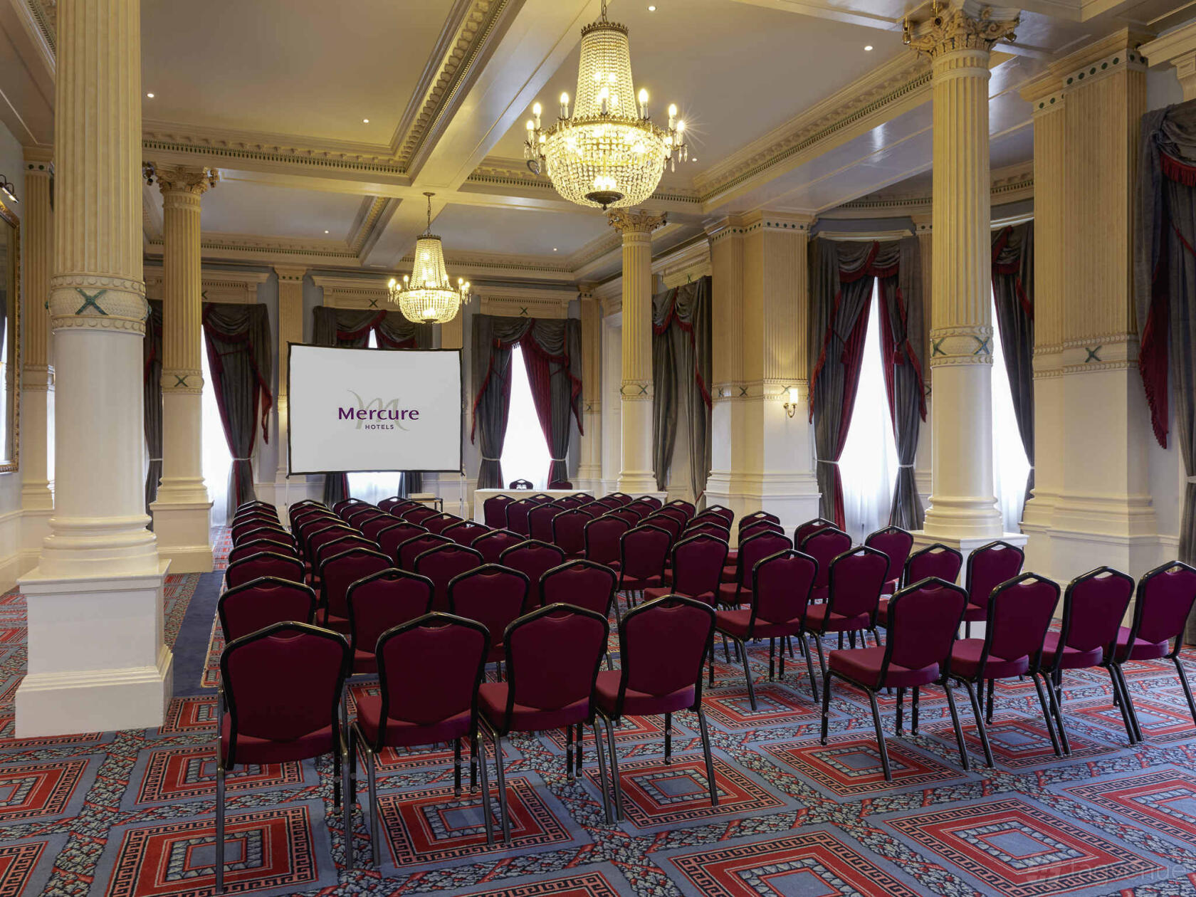 A ballroom with chandeliers, tall ornate columns, and rows of red chairs facing a projector screen at Mercure Exeter Rougemont Hotel.