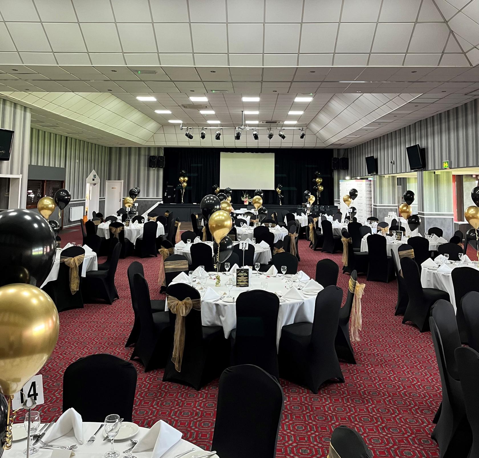 An event venue with round tables, black and gold balloons, and a stage with a projector at Walsall Football Club.