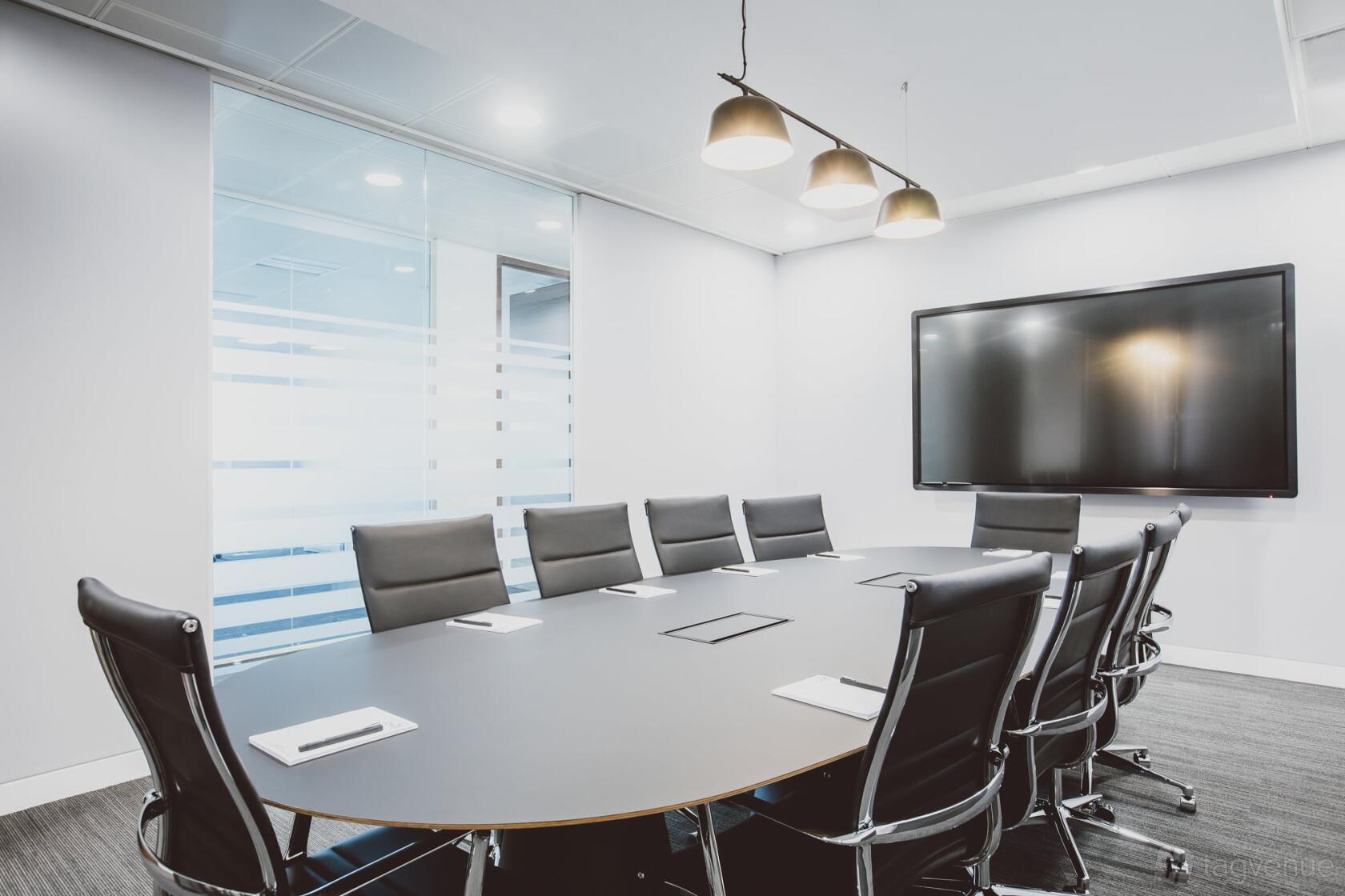 A meeting room with a large oval table, black leather chairs, a wall-mounted screen, and frosted glass walls at Orega Uxbridge.