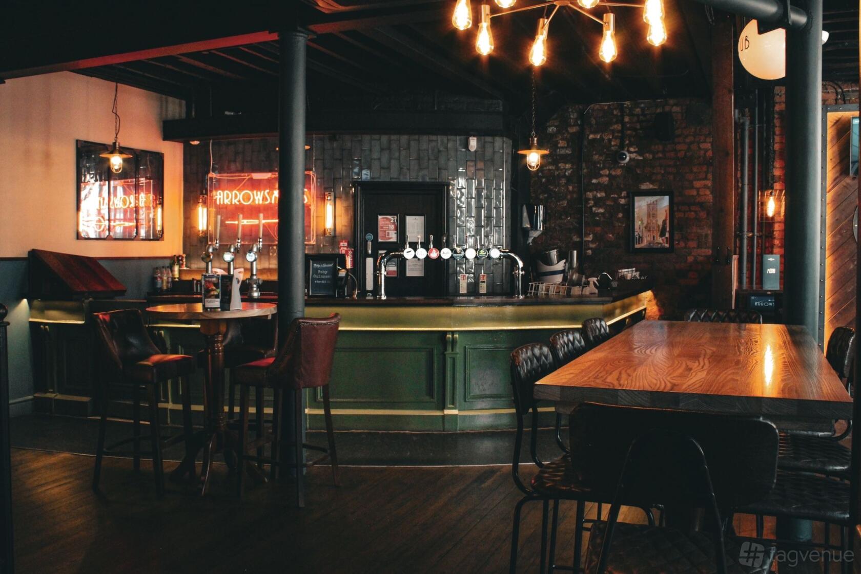 An event space in a pub with a green bar, exposed brick walls, industrial lighting, and wooden tables at Arrowsmiths @ The Shipping Forecast Liverpool.