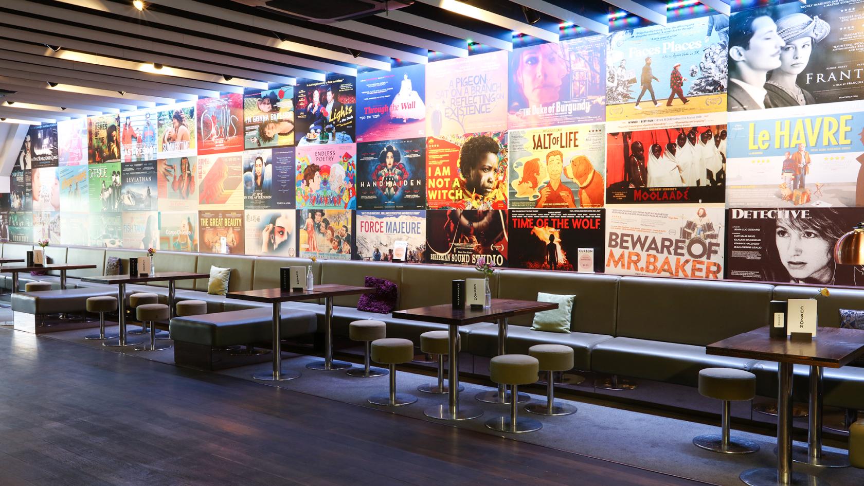 A cinema bar with film poster wall decor, cushioned bench seating, and small round tables at Curzon Wimbledon.