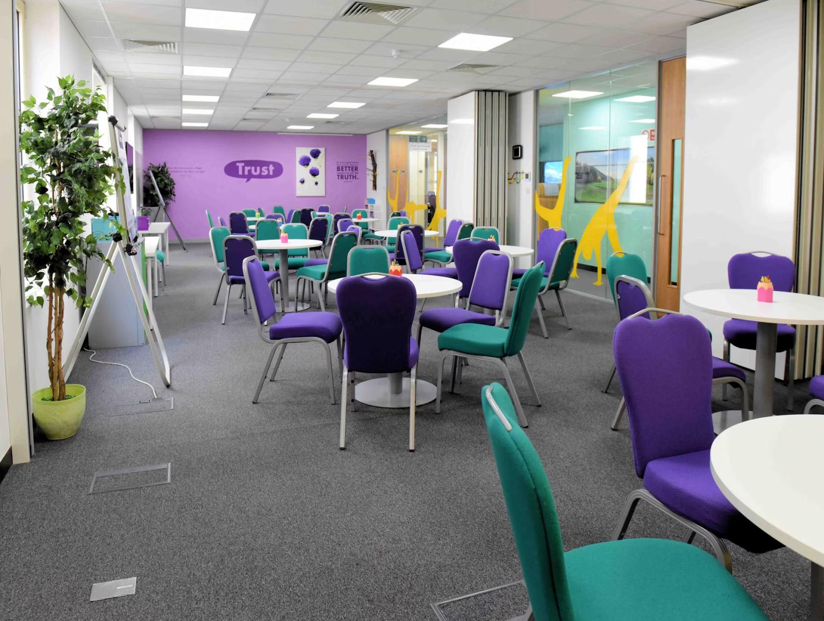 A meeting room with round tables, purple and teal chairs, carpeting, and a wall with motivational text at Happy.