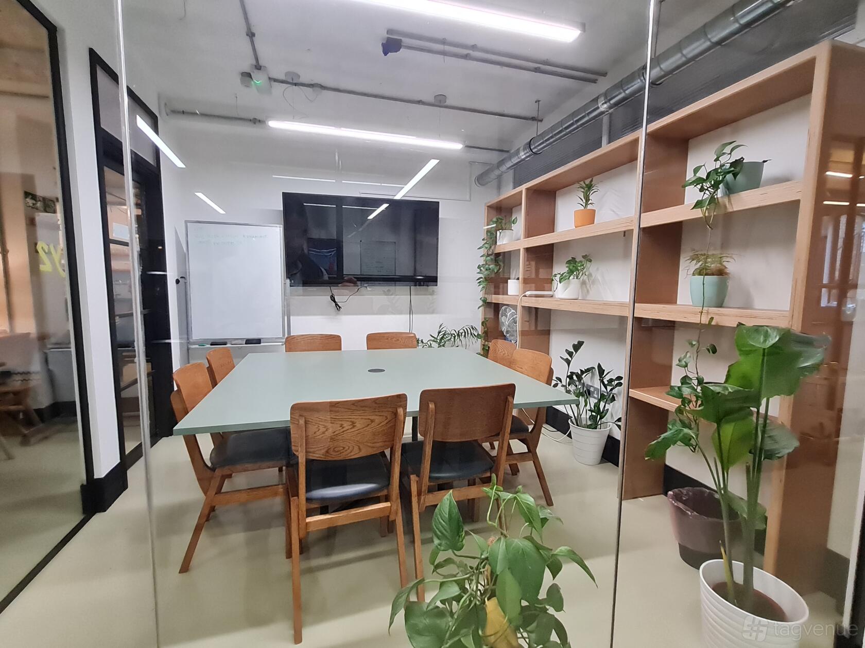 A meeting room with a glass wall, wooden table and chairs, potted plants, and a wall-mounted screen at The Worker's League.