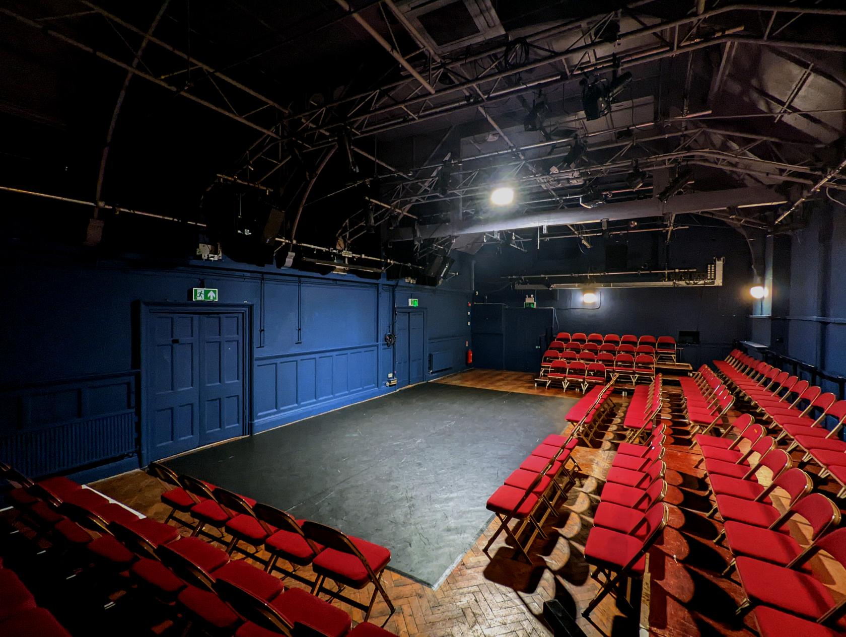 A theatre with tiered seating, red cushioned chairs, and exposed ceiling lighting at Theatro Technis.