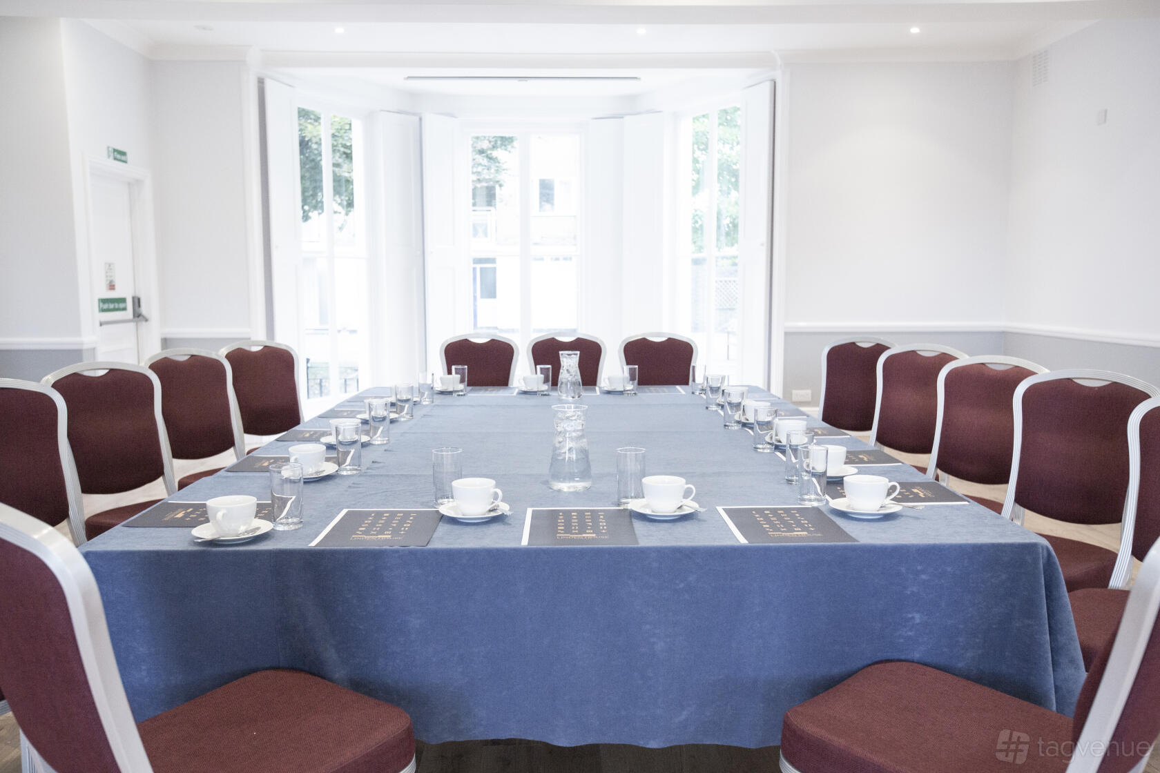A ballroom with bay windows, red upholstered chairs, and a U-shaped table set for a meeting at Linden House Riverside.