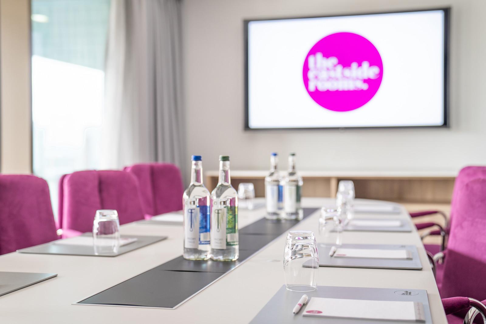 A boardroom with magenta chairs, bottled water, glasses, and a large screen at The Eastside Rooms.