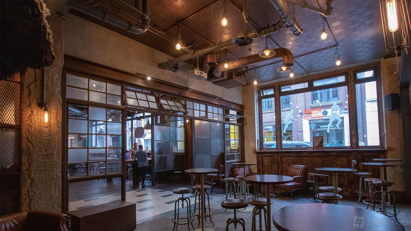 A bar in a pub with industrial-style exposed pipes, tall windows, and round wooden tables at Long Arm Brewery & Tap Room.