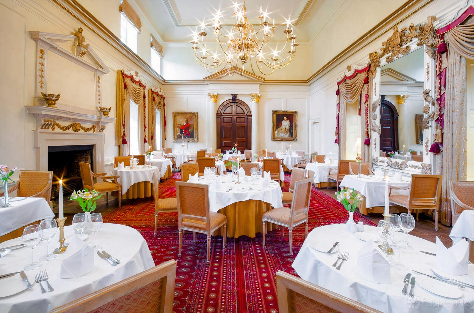 An event space with arched windows, ornate chandeliers, and round dining tables with white linens at Hintlesham Hall Hotel.