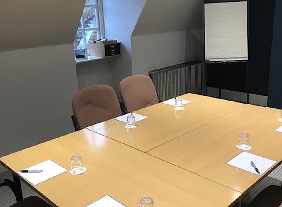 A meeting room with a rectangular wood table, beige chairs, notepads, glasses, and a flip chart at Barnett Hill Hotel.