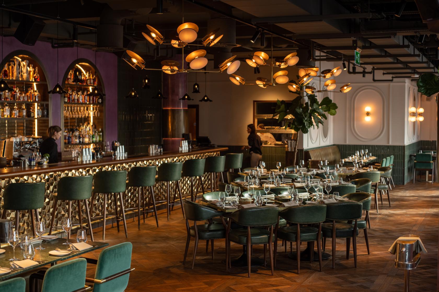 A rooftop restaurant with velvet chairs, round tables set for dining, and a long bar with modern lighting at Wagtail.