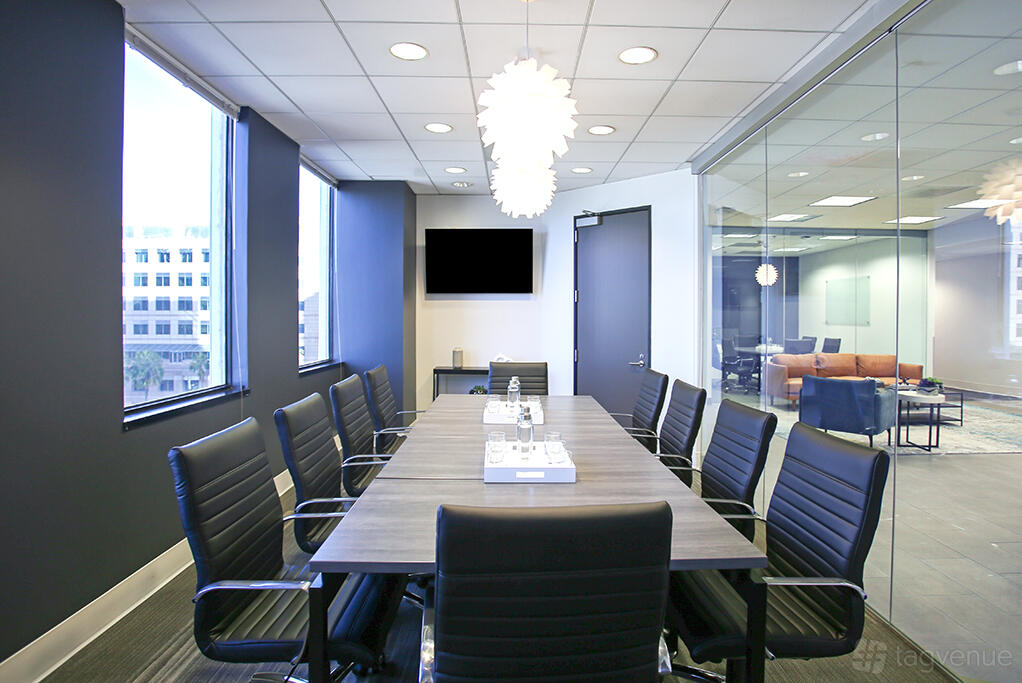A meeting room with a long conference table, black leather chairs, large window, and pendant lighting at Premier Workspaces - 444 W Ocean.
