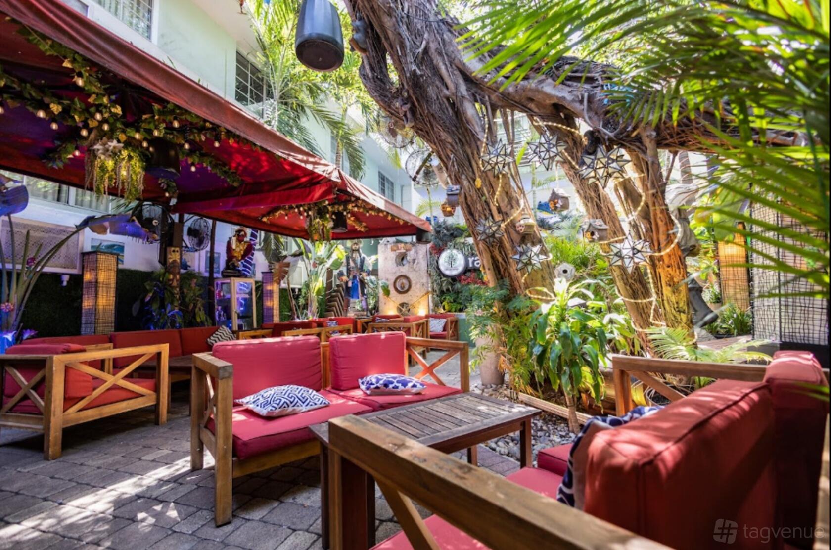 An outdoor restaurant patio with red cushioned seating, potted plants, and large trees at Rakija Lounge.