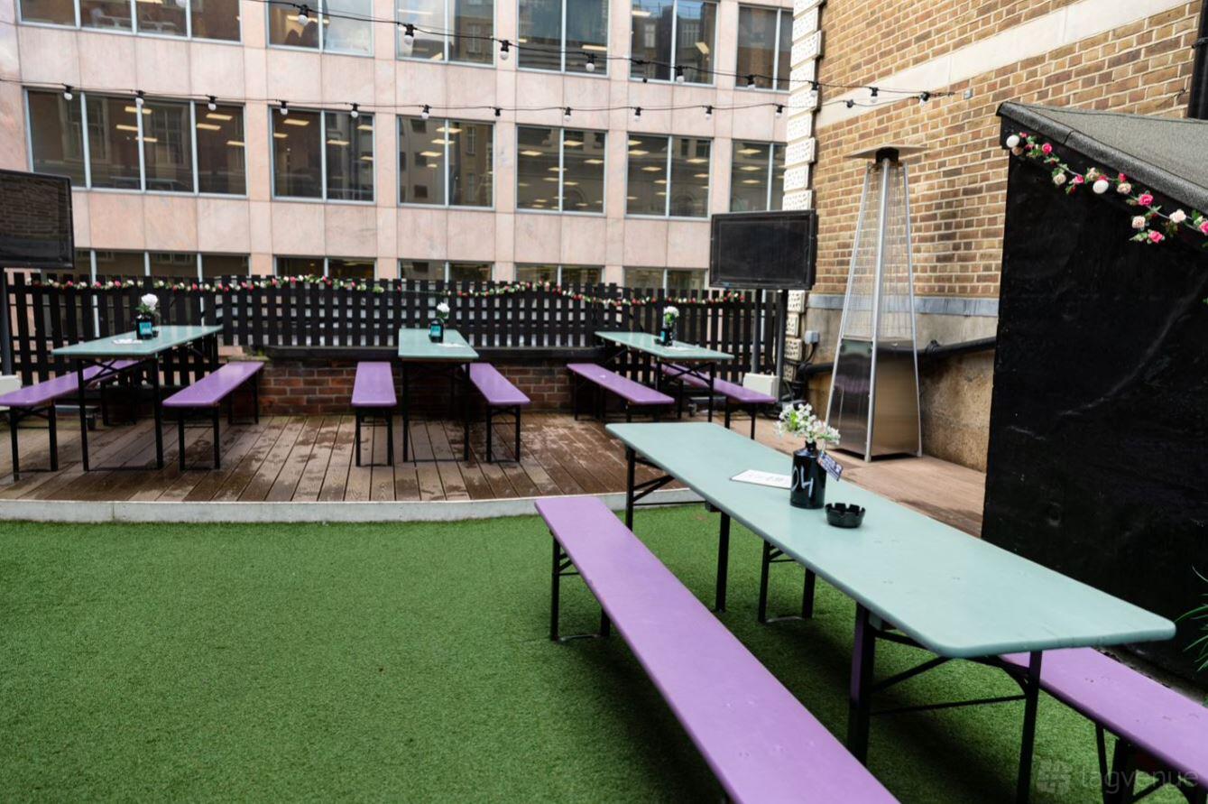 A terrace at a pub with picnic tables, artificial grass, flowers, and string lights at Three Tuns Aldgate.