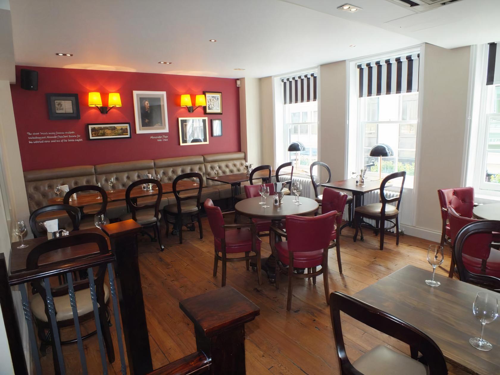 A pub dining room with wooden floors, red upholstered chairs, framed art, and wall sconces at The Clarence.