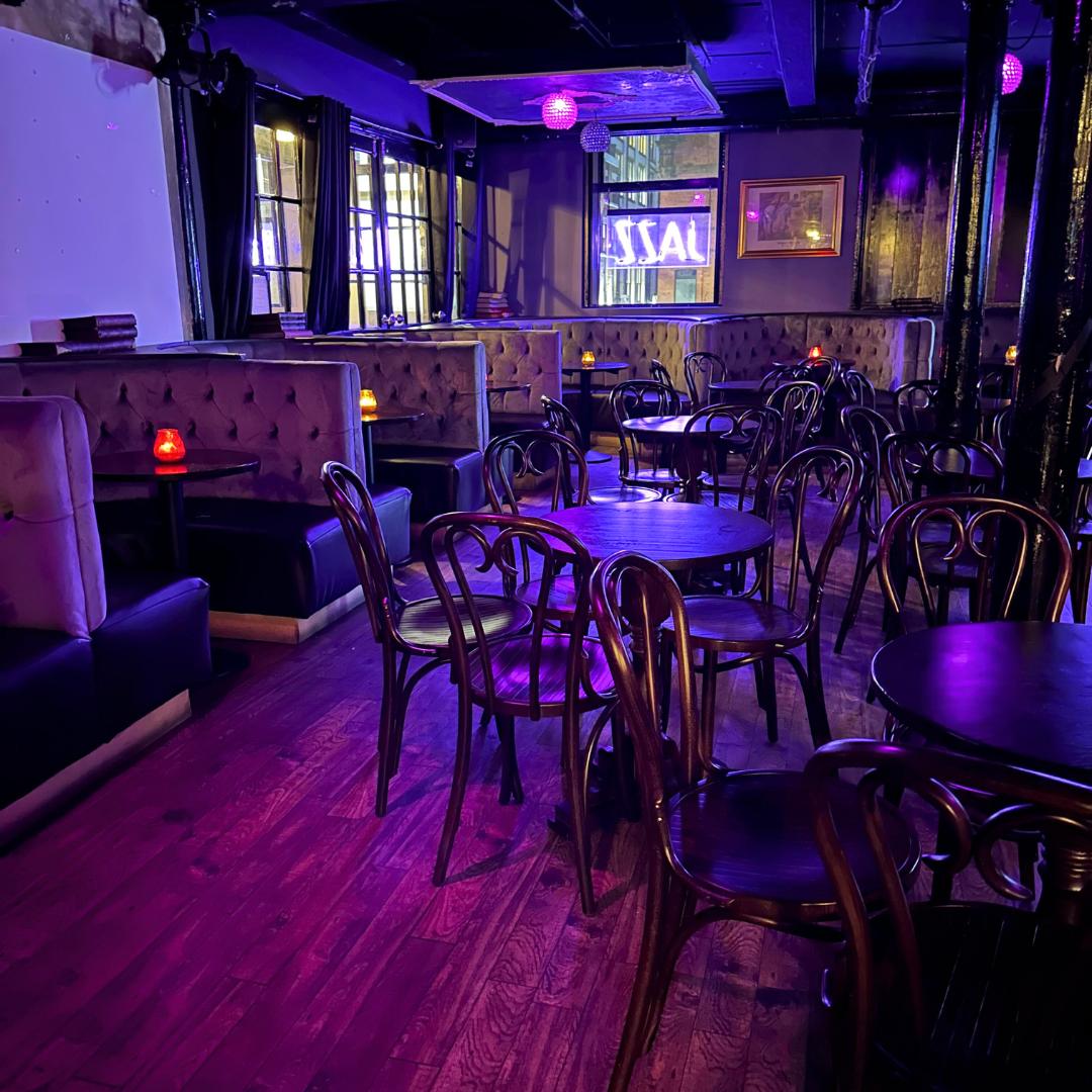 A bar with tufted booth seating, round dark wood tables, and purple ambient lighting at The Fitzgerald.