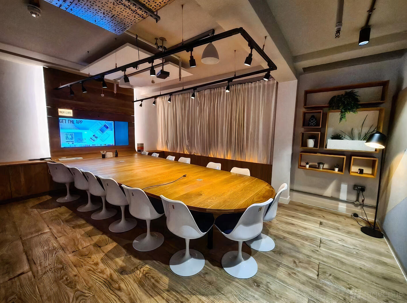 A meeting room with a long wooden table, white chairs, mounted screen, and wall shelving at Huckletree Soho.