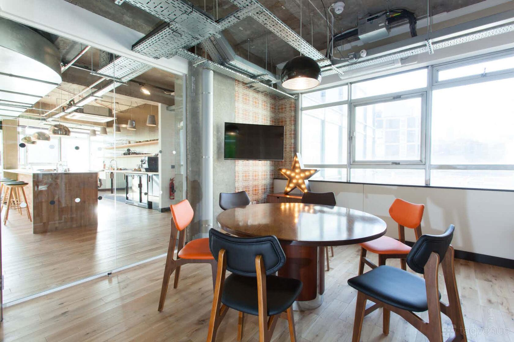 A meeting room with a round wooden table, colorful chairs, large windows, and exposed ceiling at The Trampery Old Street.