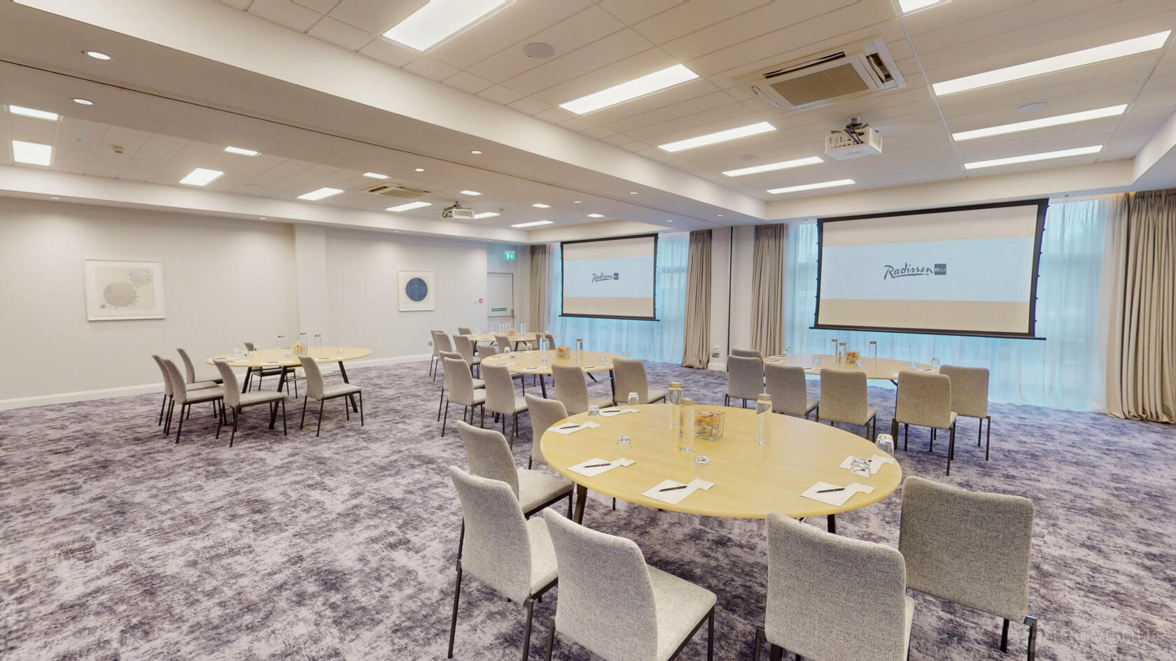A meeting room with round tables, grey chairs, large projection screens, and natural light at Radisson Blu Hotel, Dublin Airport.