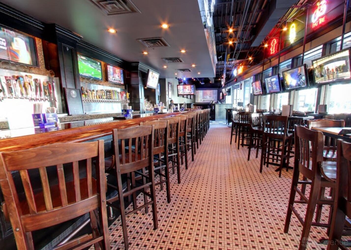 A bar with a long wooden counter, multiple barstools, wall-mounted taps, and flat screen TVs at Hudson Grille - Downtown.