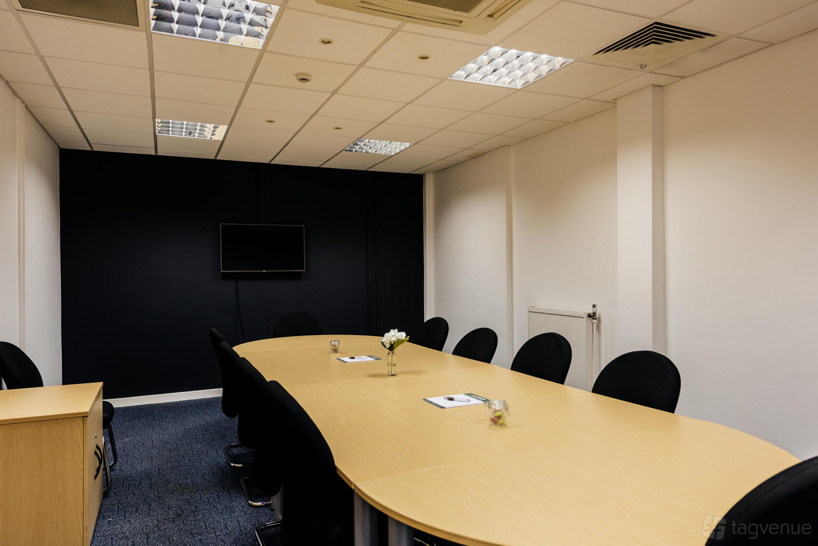A meeting room with an oval conference table, black chairs, a wall-mounted screen, and a vase at Melton Court.
