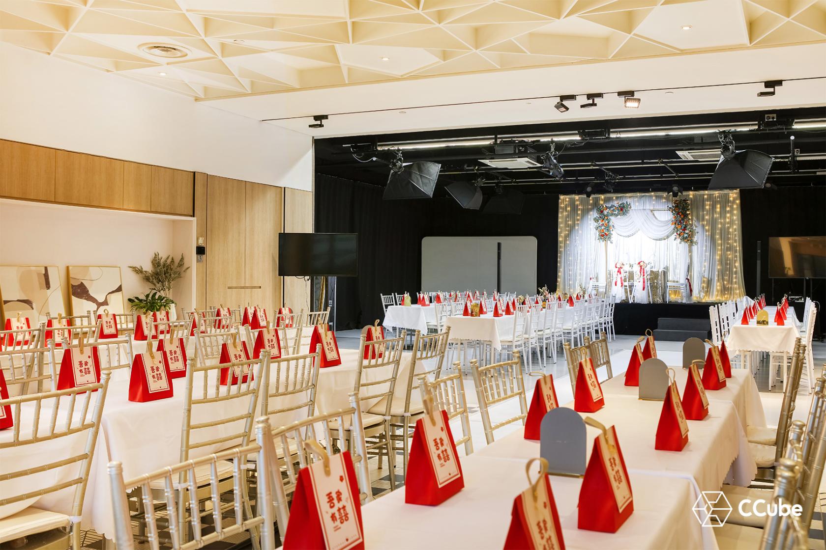 An event venue with rows of white chiavari chairs, banquet tables with red gift bags, and a stage at CCube Studio Weave.