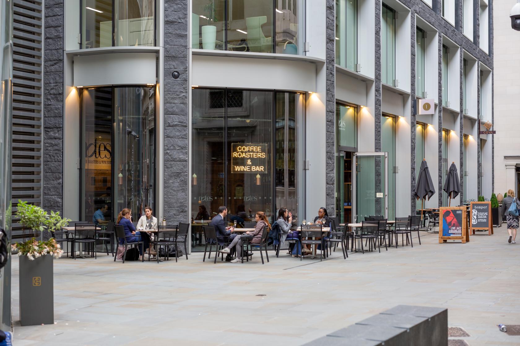 A cafe with outdoor seating, glass walls, and modern stone facade at Notes Coffee & Bars - Bank.