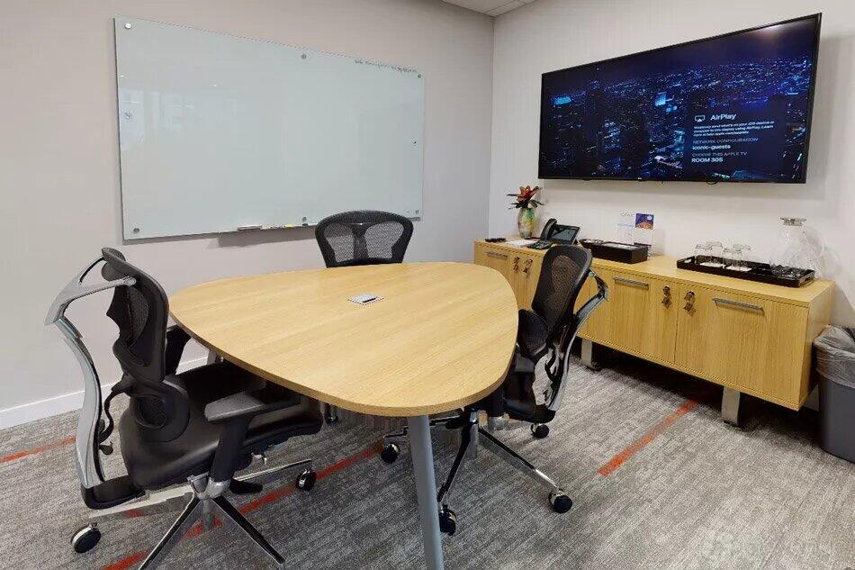 A meeting room with a triangular wooden table, four mesh chairs, a wall-mounted screen, and a whiteboard at Iconic Workspaces @ Aventura.