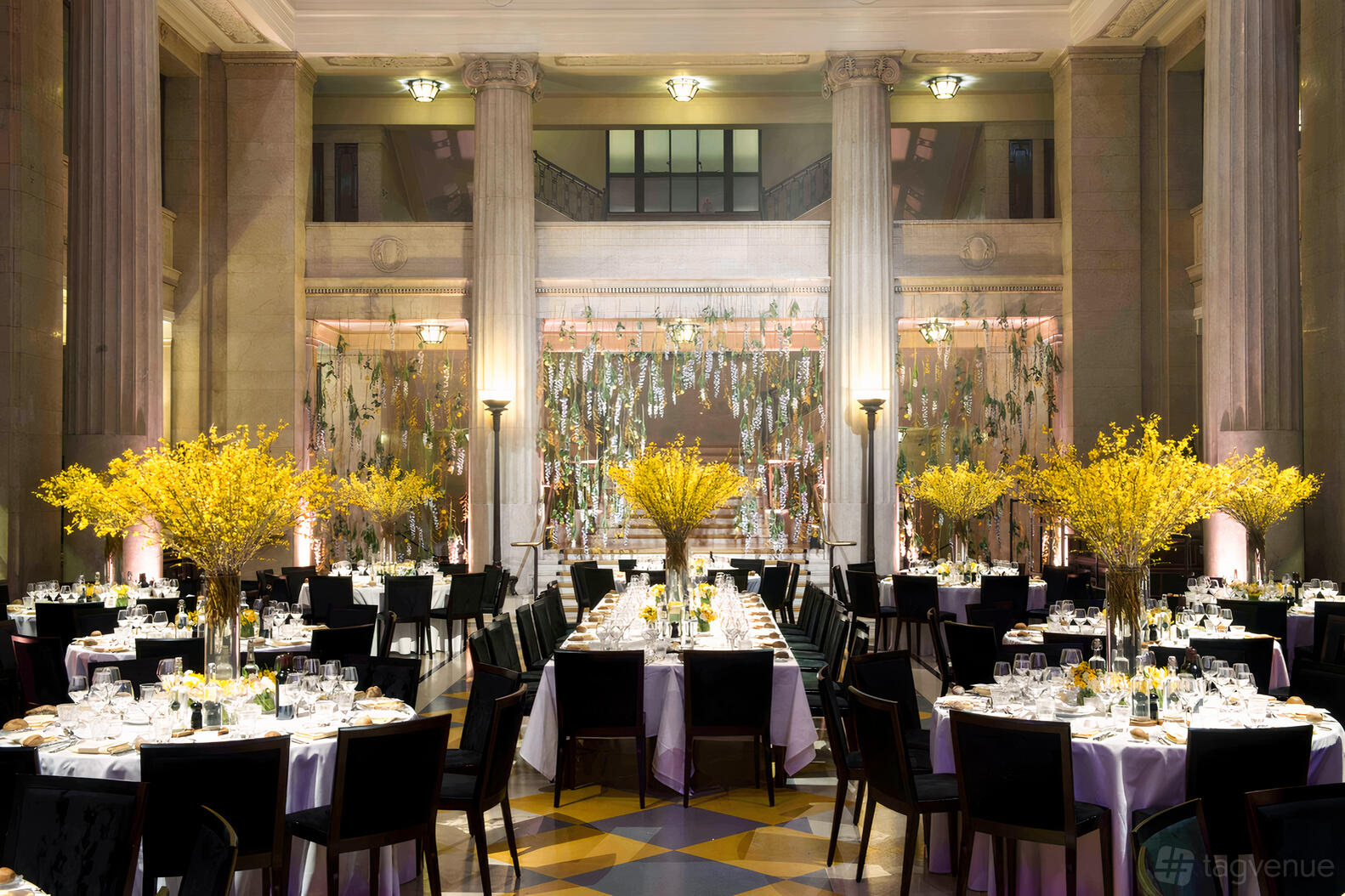 A grand hall with marble columns, high ceilings, round tables set with white linens and yellow floral centerpieces at Banking Hall.