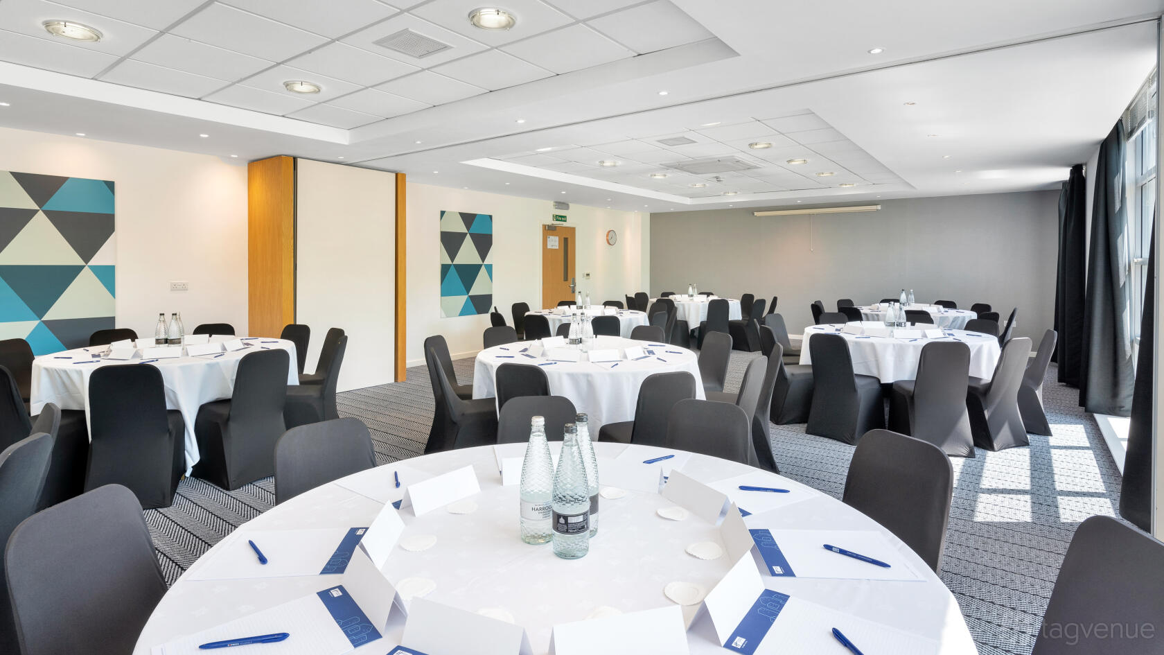 An event space with round tables, white linens, and bottled water setup in the Pennington Suite at Holiday Inn Express Leigh Sports Village.