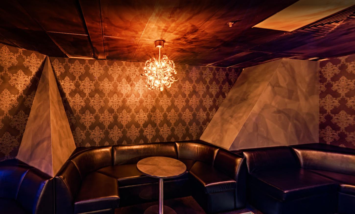 A nightclub lounge with patterned wallpaper, a black leather sectional sofa, and a chandelier at Cocktails Nightclub.