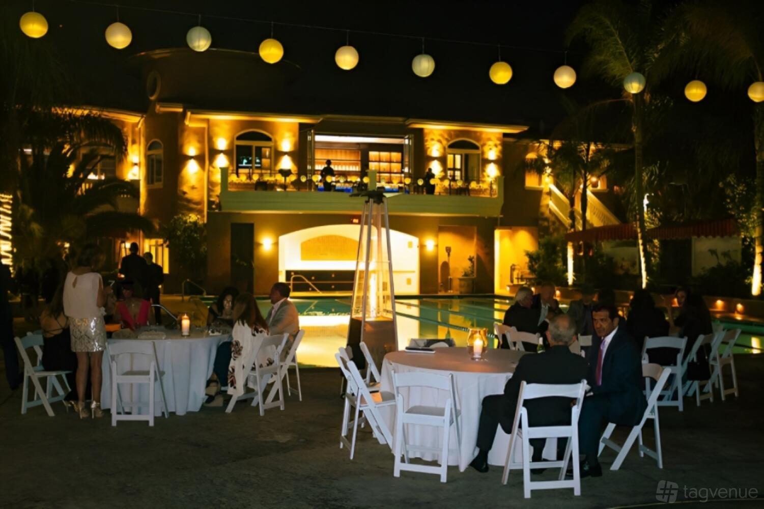 An outdoor pool area with round tables, white chairs, string lights, and palm trees at South End Racquet And Health Club.
