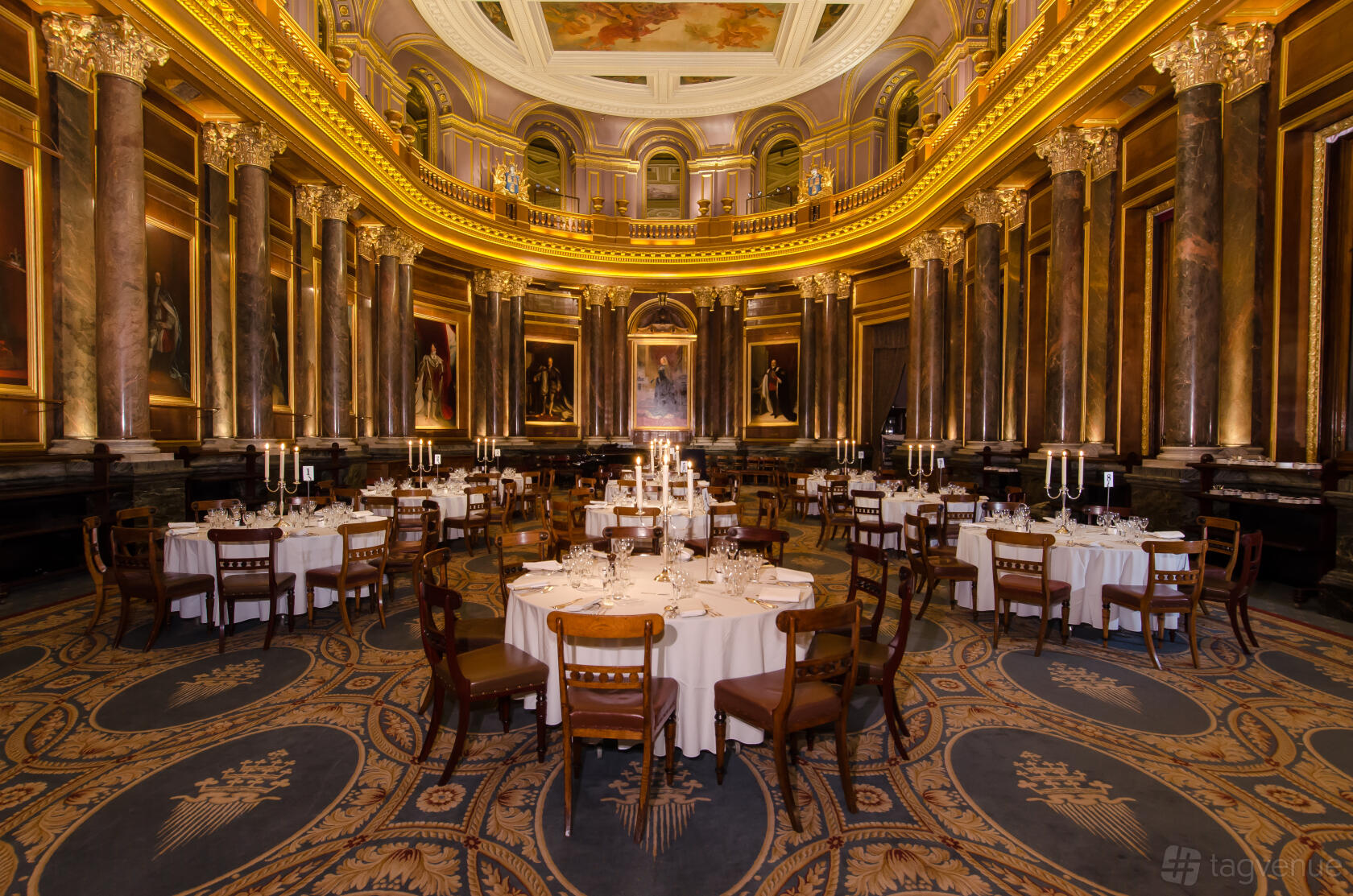 A grand hall with marble columns, ornate gold trim, chandeliers, and round tables with white linens at Drapers' Hall.
