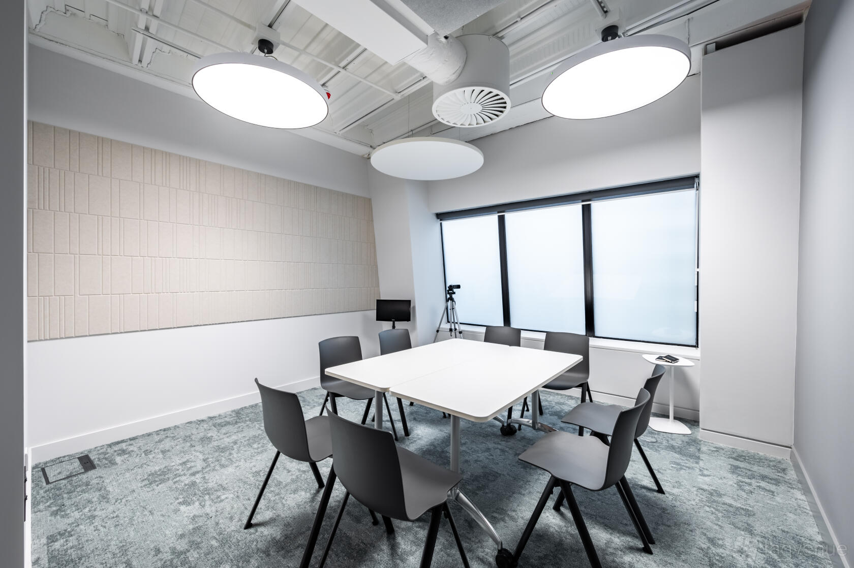 A meeting centre with a square table, eight chairs, large round ceiling lights, and a window at Spotlight Studios.
