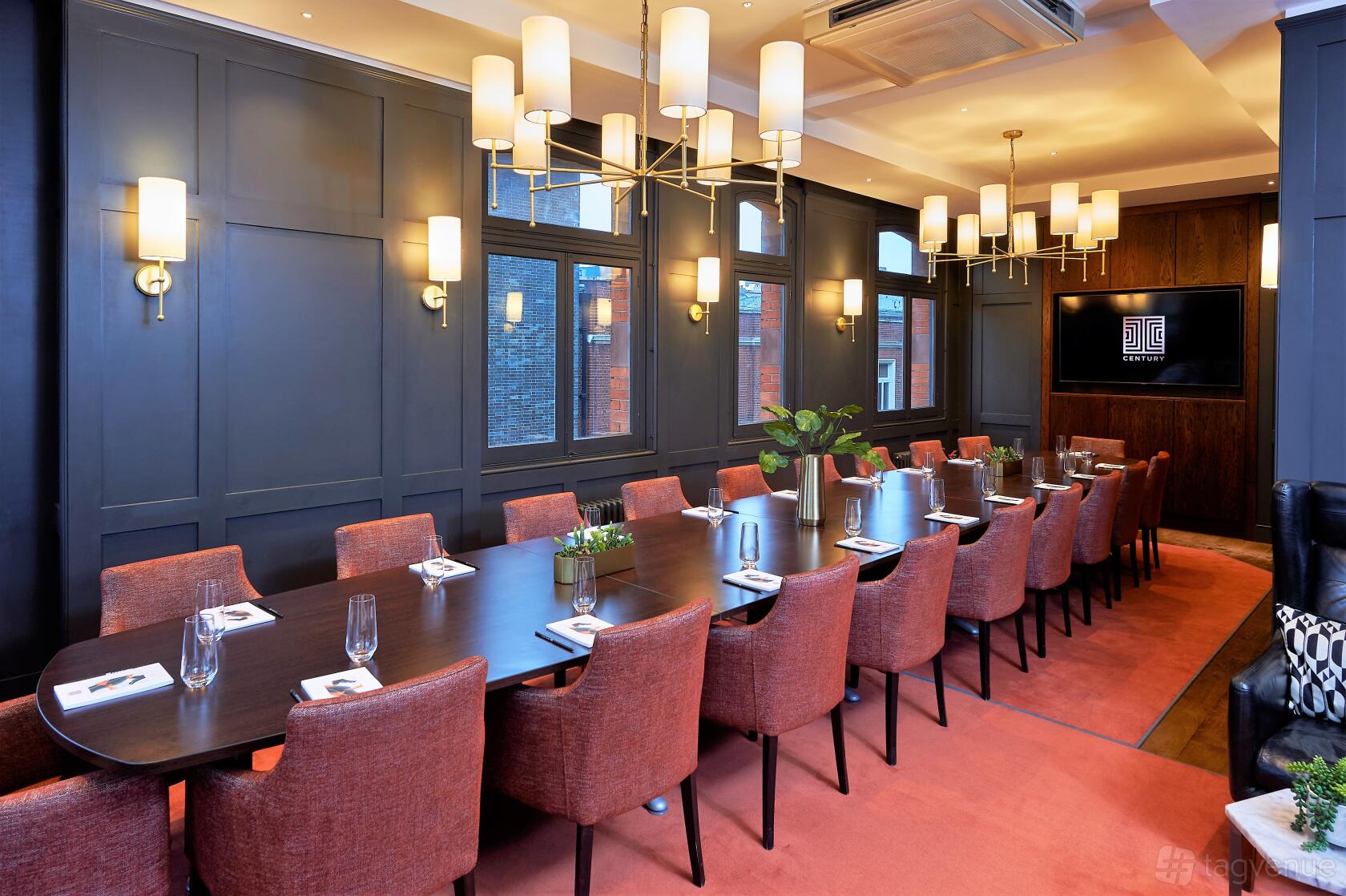 A private bar room with a long dark wood table, red upholstered chairs, pendant lighting, and navy paneled walls at Century Club.