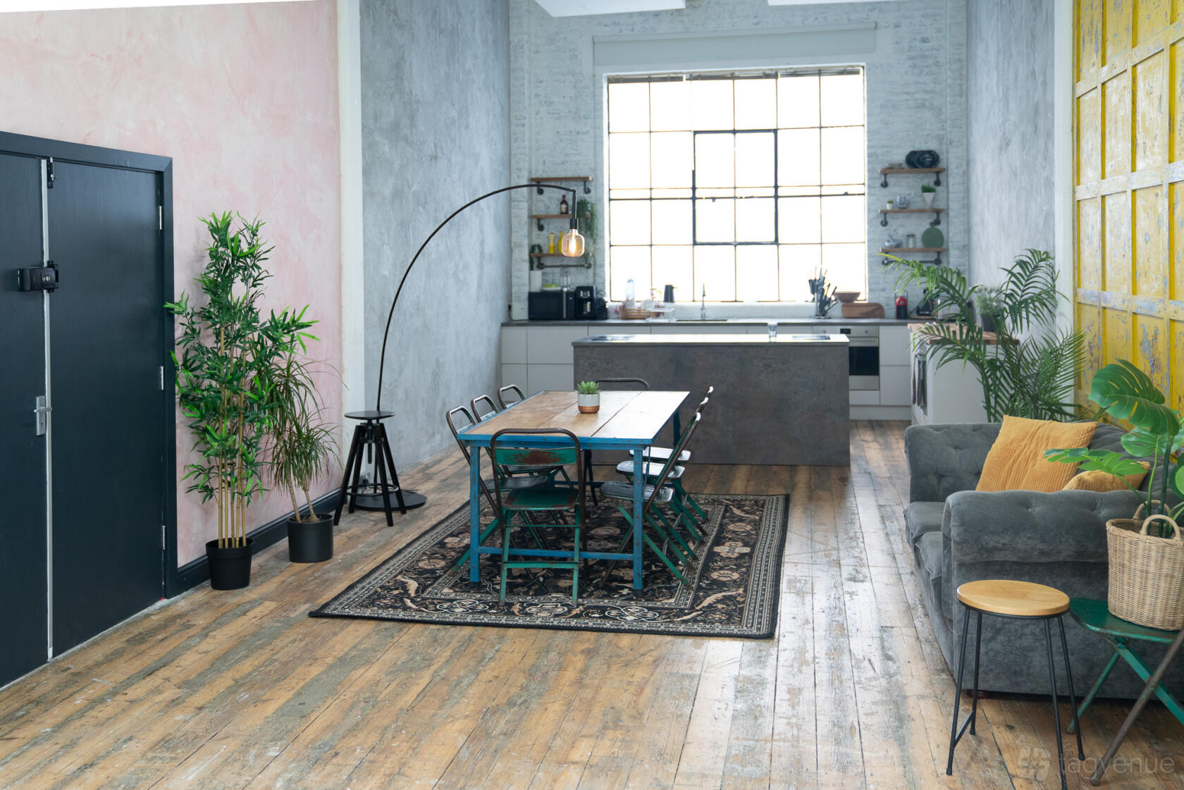 A photo studio with a large industrial window, wood floors, kitchen island, and potted plants at Crixus Studios.