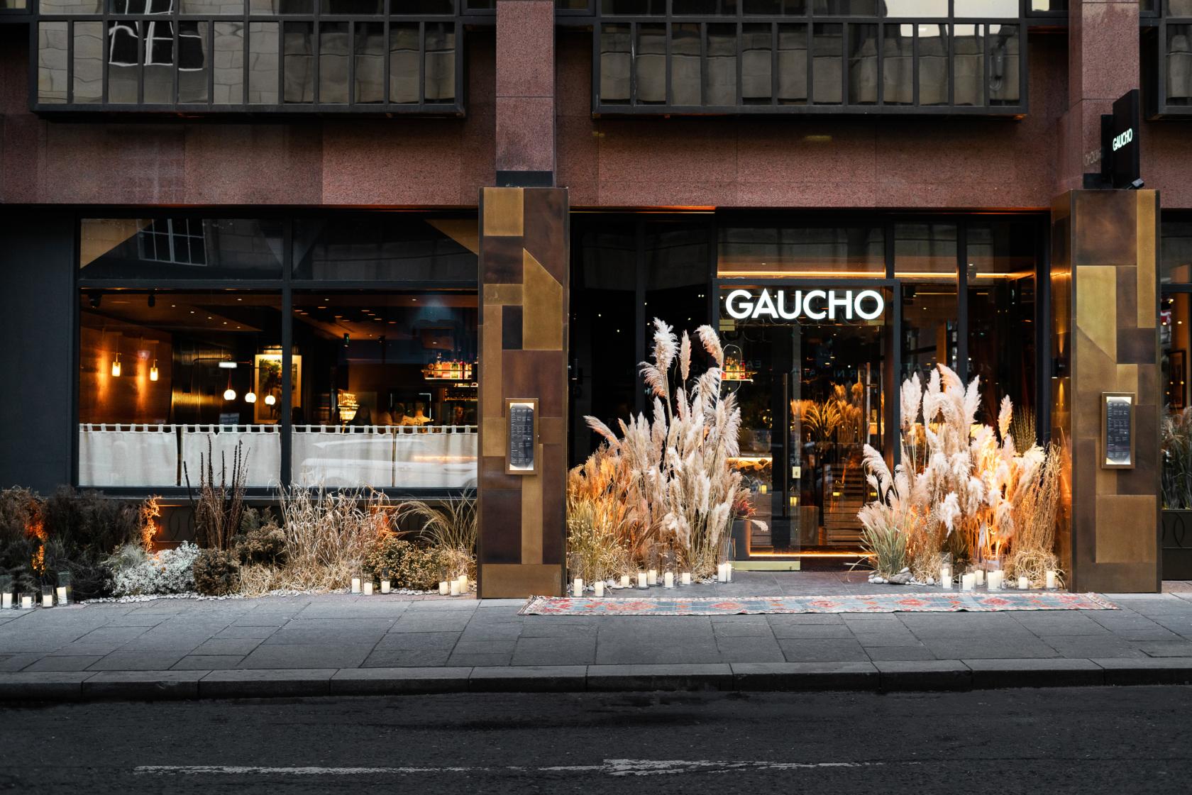 A restaurant entrance with tall glass windows, illuminated signage, and pampas grass arrangements at Gaucho Glasgow.
