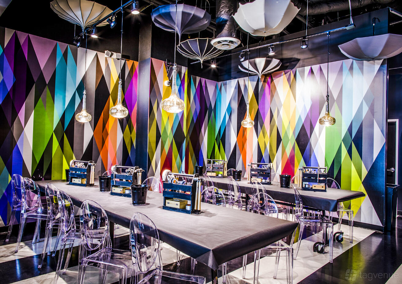 A bar with geometric multicolored walls, long tables with black tablecloths, and clear chairs at Upstairs Circus ATX.