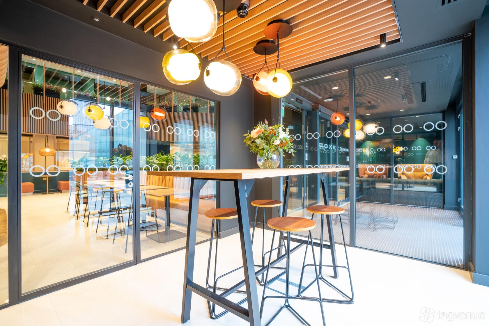 A meeting room with glass walls, round pendant lights, and high tables with stools at Orega Balloon Street.