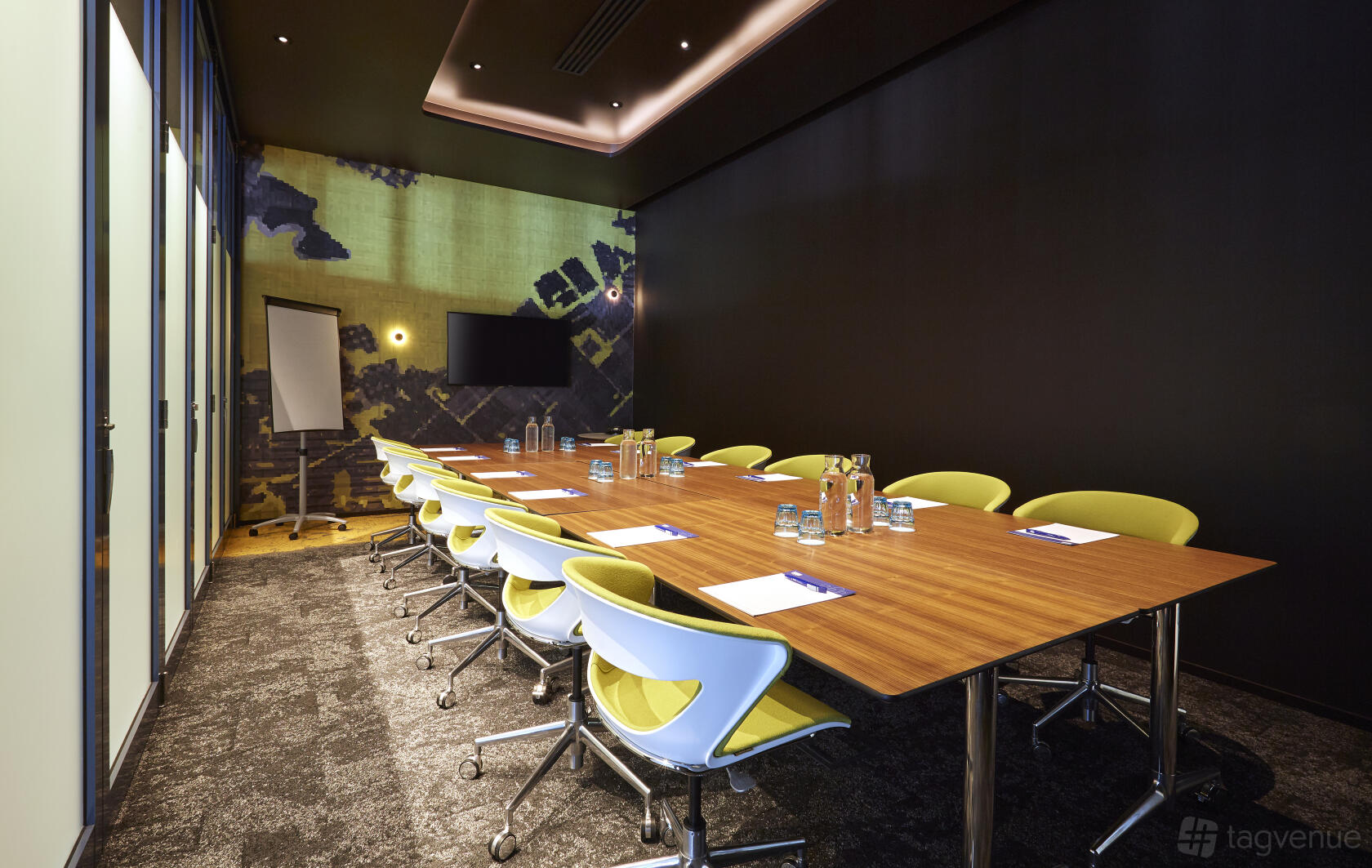 A meeting room with a long wooden table, yellow chairs, glass water bottles, and a wall-mounted screen at Holiday Inn Express Newcastle.