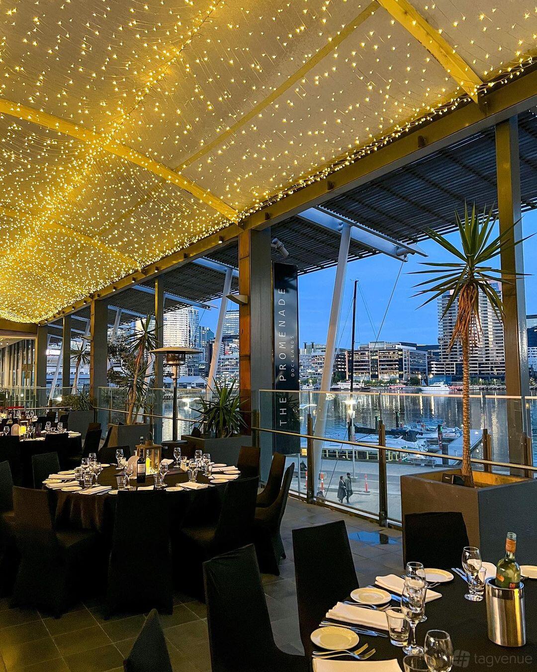 A hall with floor-to-ceiling windows, waterfront views, and a ceiling draped in fairy lights at The Promenade Docklands.