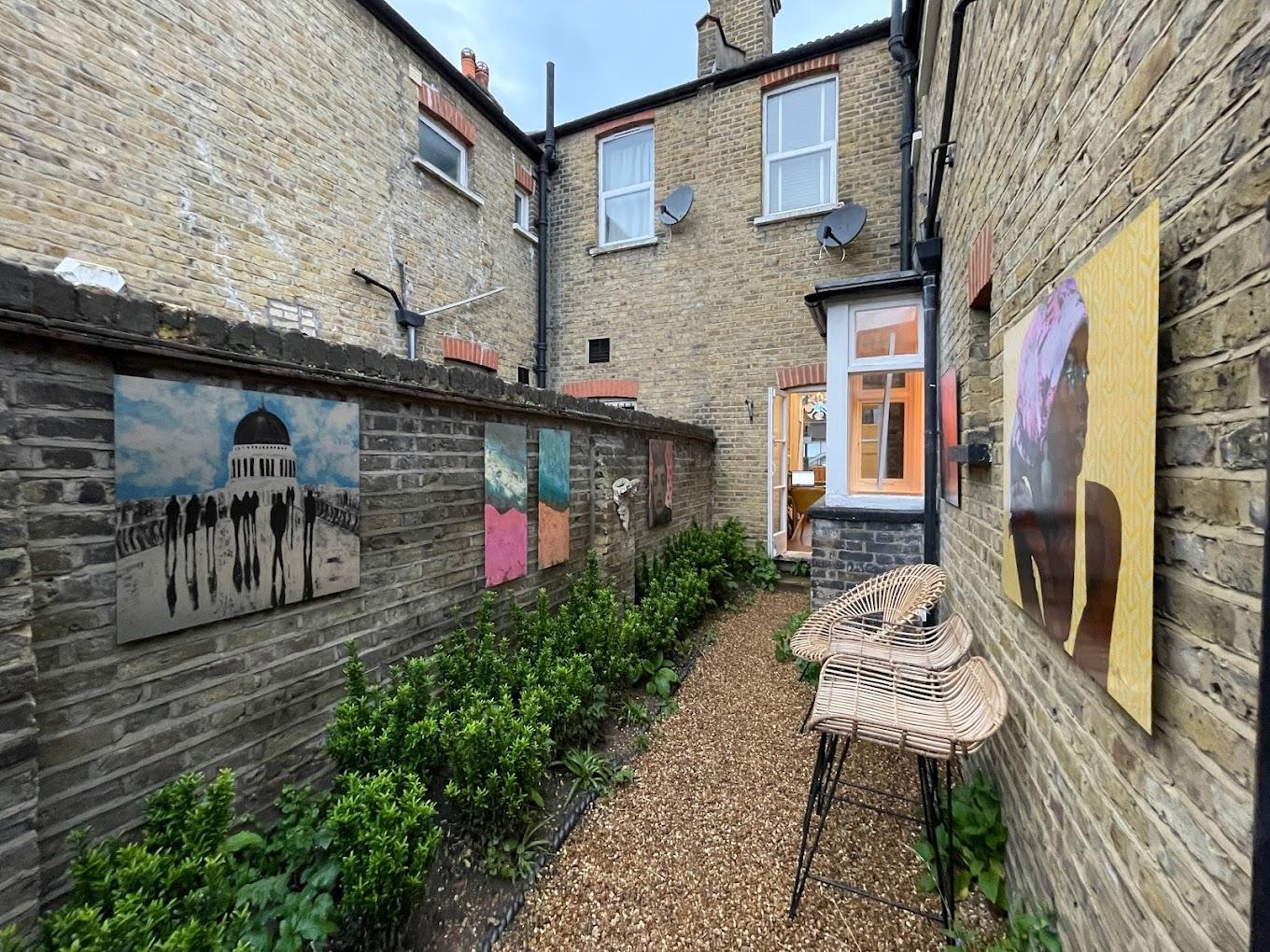 An outdoor gallery with artwork displayed on brick walls, greenery, and wicker chairs at Ad Lib Gallery, 10 Church Road.