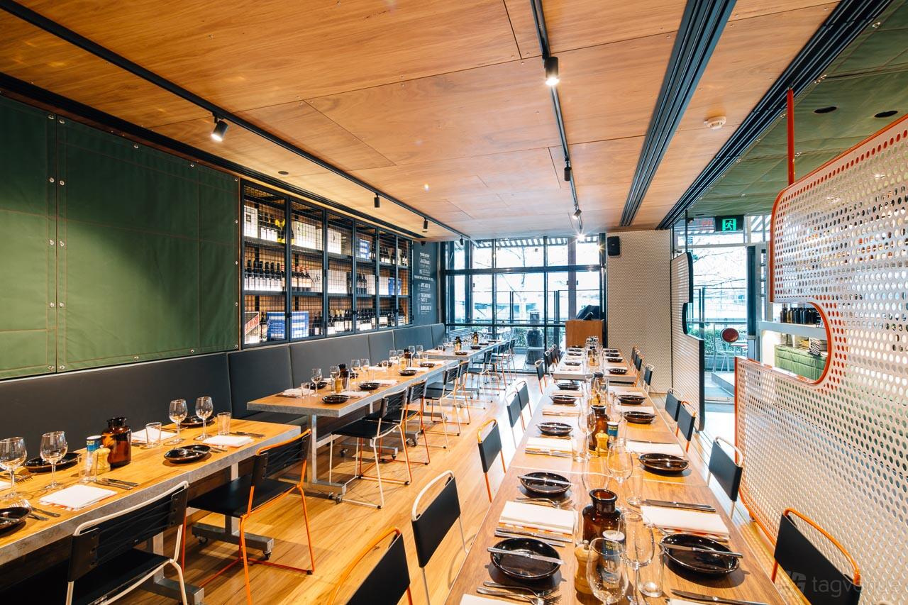 A restaurant with floor-to-ceiling windows, long wooden tables set with black dinnerware, and exposed shelving at BearBrass.