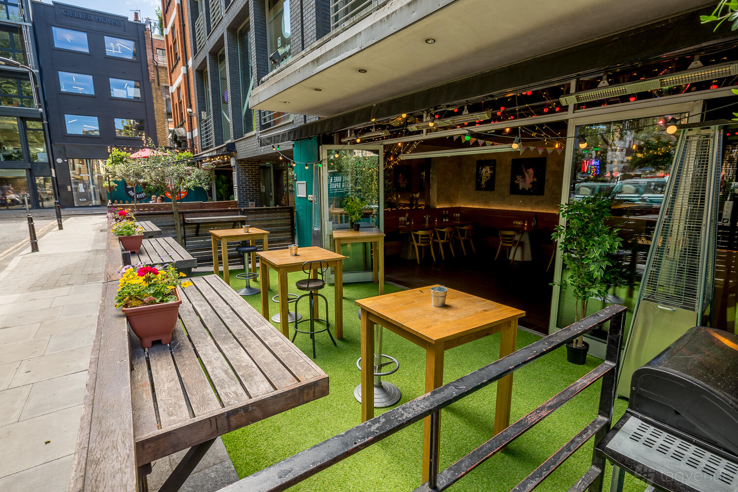 A bar with outdoor seating on artificial grass, potted plants, and open sliding doors at Colours Hoxton.