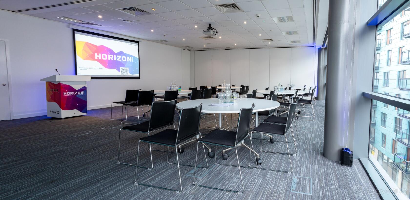 A training room with round tables, black chairs, bottled water, and a projector screen at Horizon Leeds.