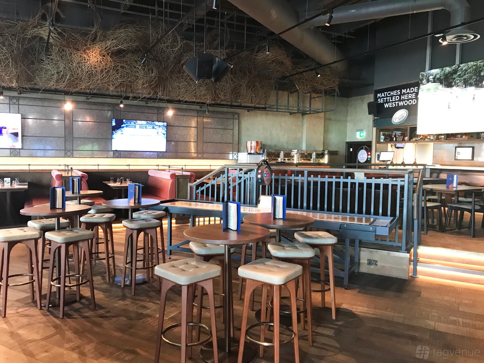 A bar with high tables, cushioned stools, wall-mounted TVs, and overhead branches at Westwood.